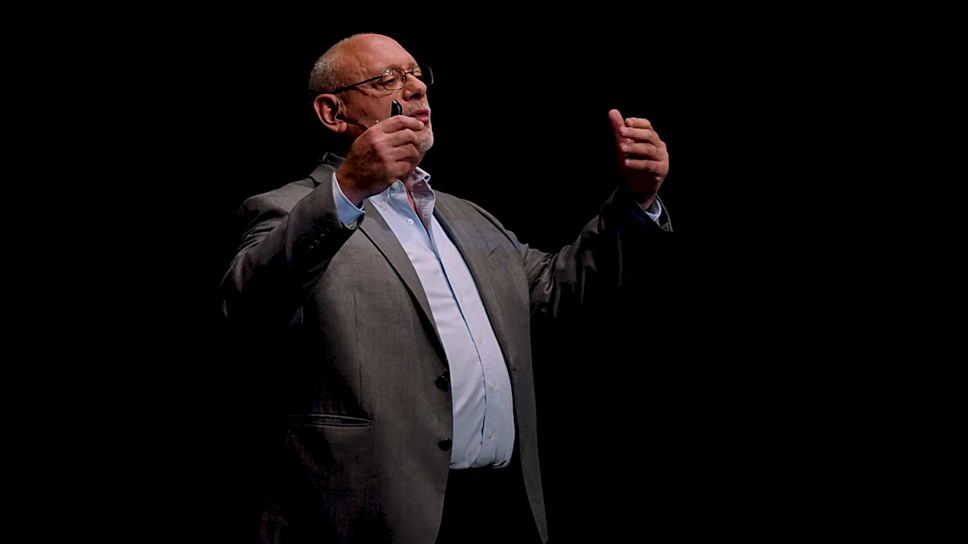 Bill Schuhlein on a dark stage, hands open, mid-explanation